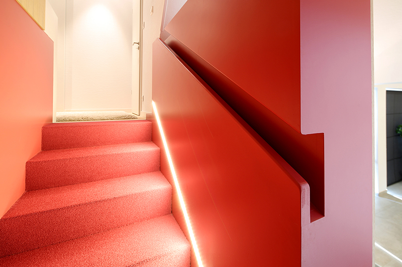 Escalier design sur mesure réalisé par LDC Agencement – L’Arche du Bois : escalier rouge moderne à gauche, mur rouge contemporain à droite avec éclairage intégré en bas, conception artisanale et agencement intérieur personnalisé.