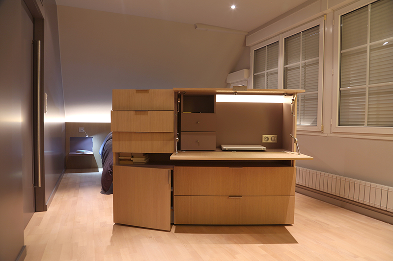 Chambre sur mesure réalisée par LDC Agencement – L’Arche du Bois : tiroirs et rangements ouverts en bois au centre de la pièce, avec trois fenêtres fermées à droite, mobilier fonctionnel et design personnalisé.