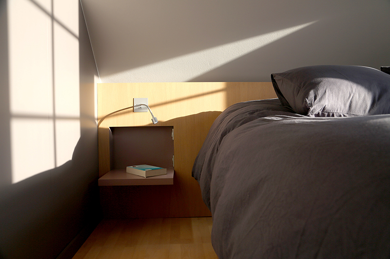 Lit gris moderne situé sur le côté droit de la pièce, intégré dans un aménagement intérieur sobre et élégant – une réalisation signée Arche du Bois.