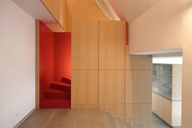 Espace intérieur conçu par Arche du Bois avec un rangement mural en bois en face, un escalier en bois en dessous et un second escalier rouge à droite, apportant un contraste audacieux dans l’aménagement