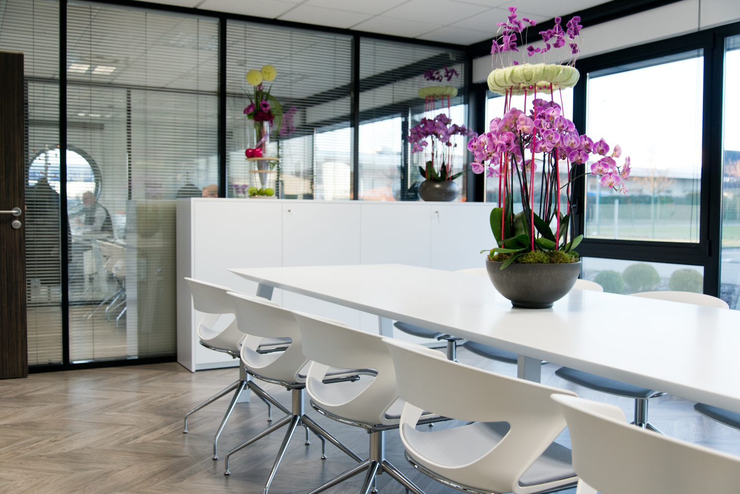 Table décorée d’un vase central, entourée de chaises disposées de part et d’autre, avec en fond une grande paroi vitrée laissant entrer la lumière naturelle — un agencement harmonieux et chaleureux signé Arche du Bois.