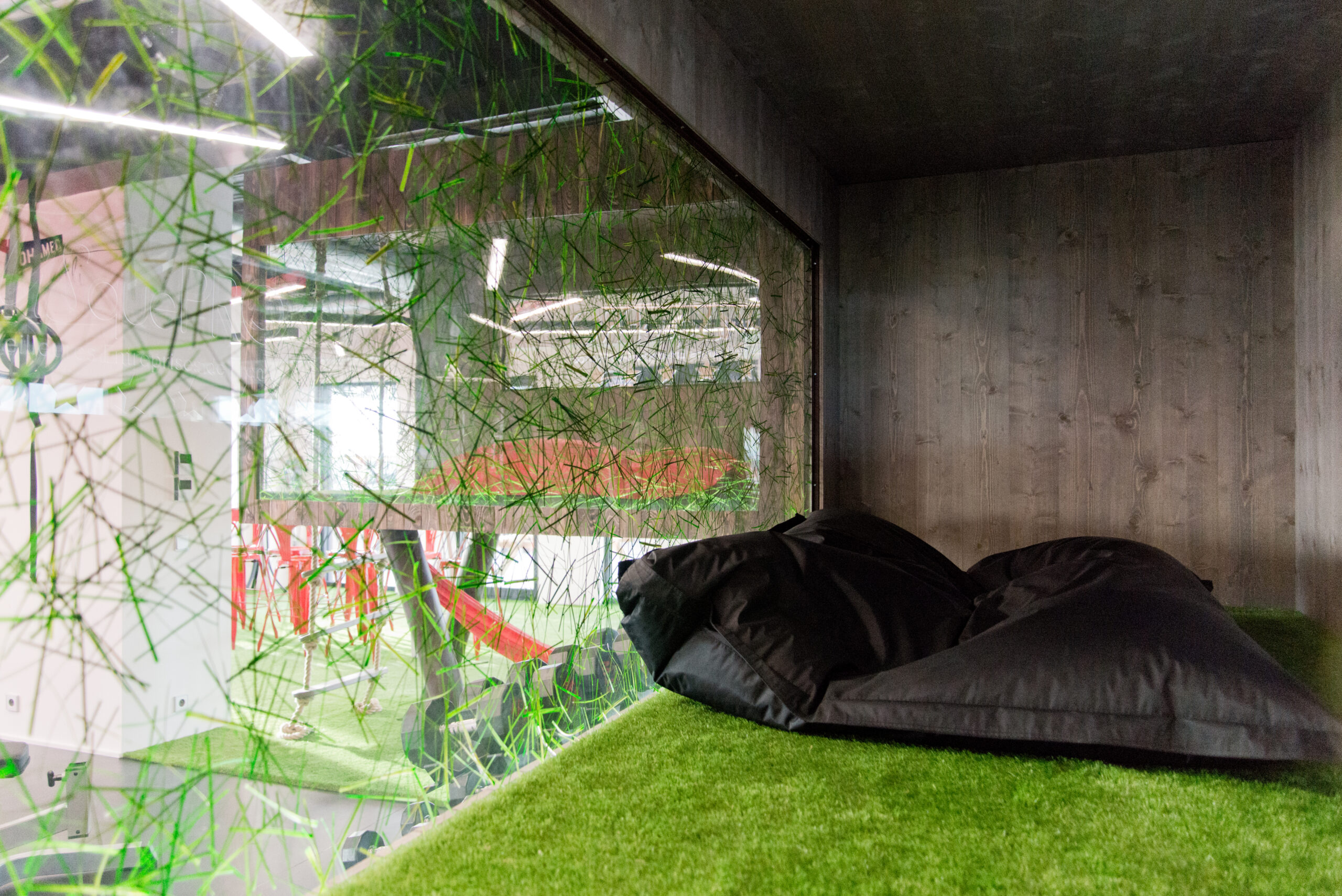 Petit espace clos avec murs en bois, paroi vitrée et sol en gazon synthétique, agrémenté d’un grand coussin de sol confortable — un agencement unique signé Arche du Bois, combinant matériaux naturels et ambiance cosy pour une zone de relaxation originale.