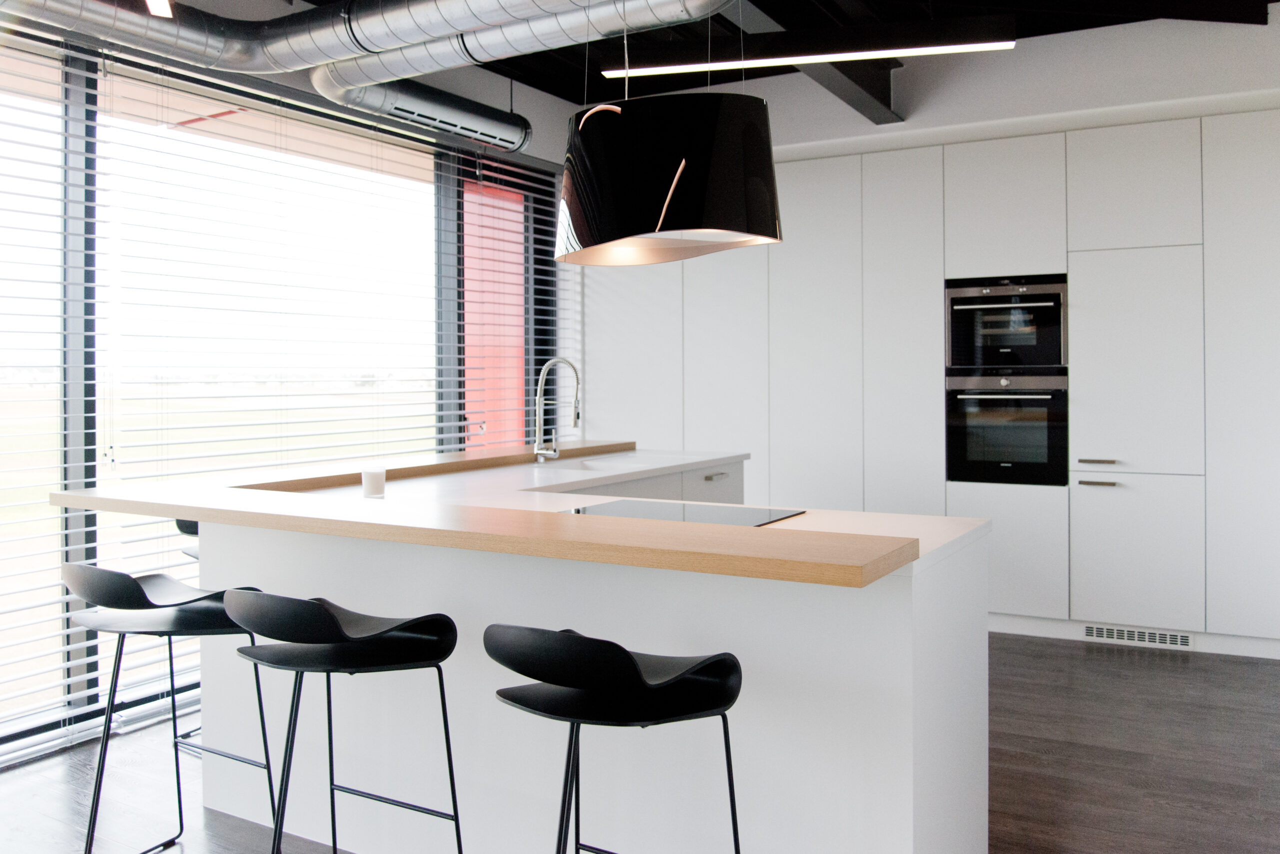 Cuisine moderne avec meubles et îlot central blancs, accompagnée de trois tabourets alignés et d’une suspension noire au-dessus du bar ; à gauche, une fenêtre habillée d’un store vénitien — un agencement lumineux et épuré signé Arche du Bois, alliant fonctionnalité et esthétique contemporaine.