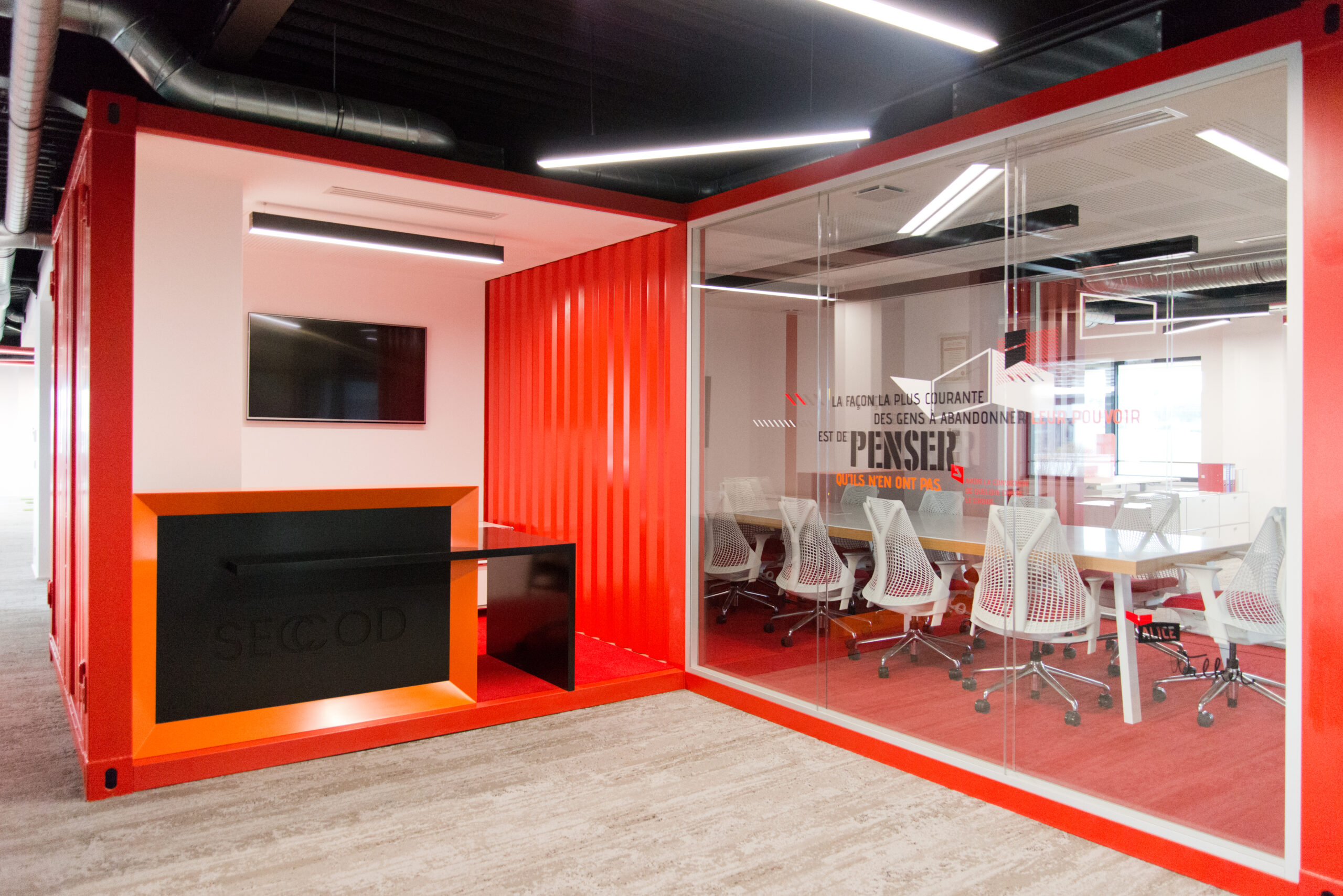 Salle de conférence avec parois vitrées teintées de rouge, équipée d’une grande table centrale et de plusieurs chaises blanches — un aménagement contemporain signé Arche du Bois, associant transparence, dynamisme et mobilier fonctionnel.