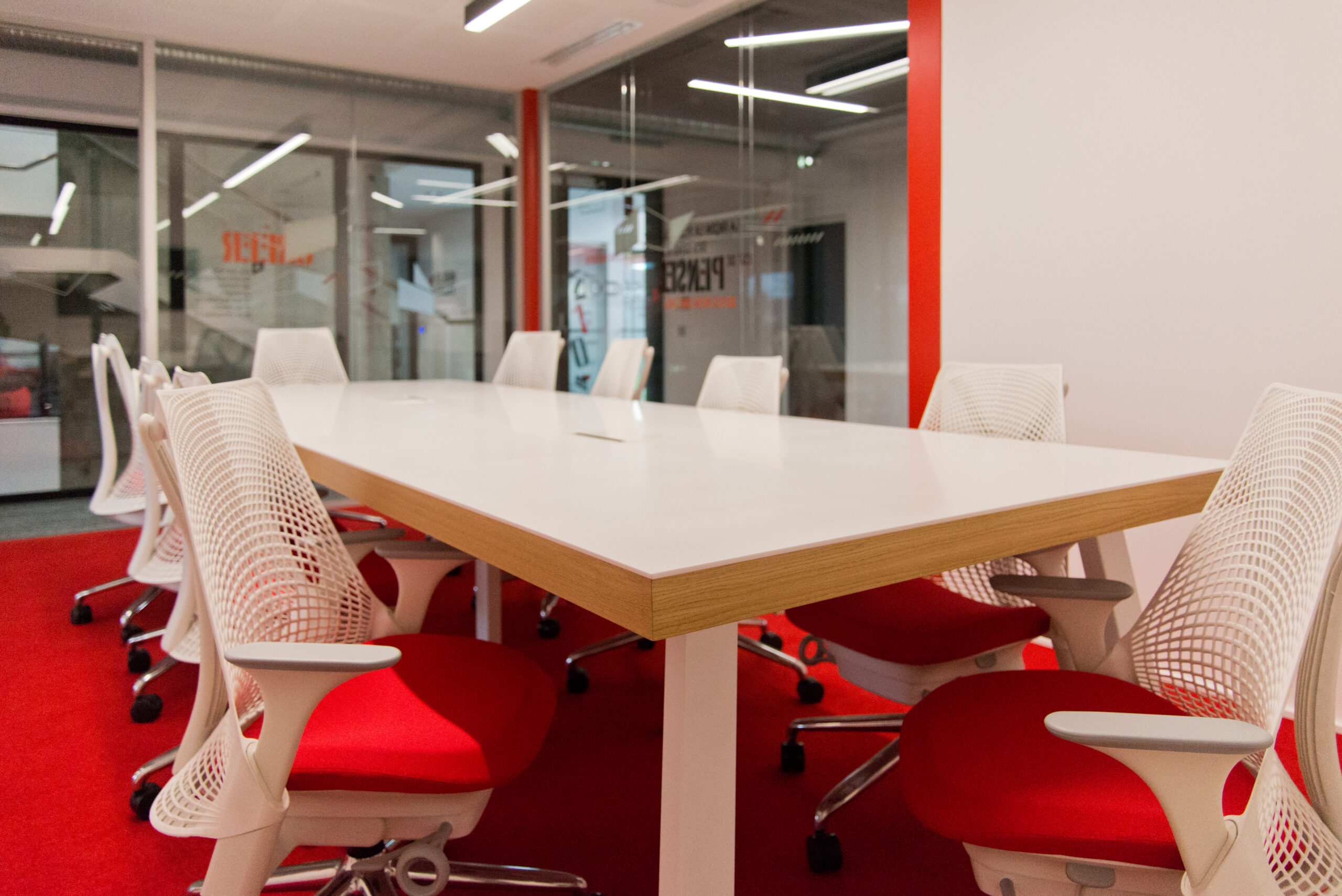 Vue intérieure d’une salle de réunion avec grande table blanche, chaises alternées rouges et blanches, et sol recouvert de moquette rouge — un aménagement dynamique et coloré signé Arche du Bois, favorisant l’échange et la créativité en milieu professionnel.