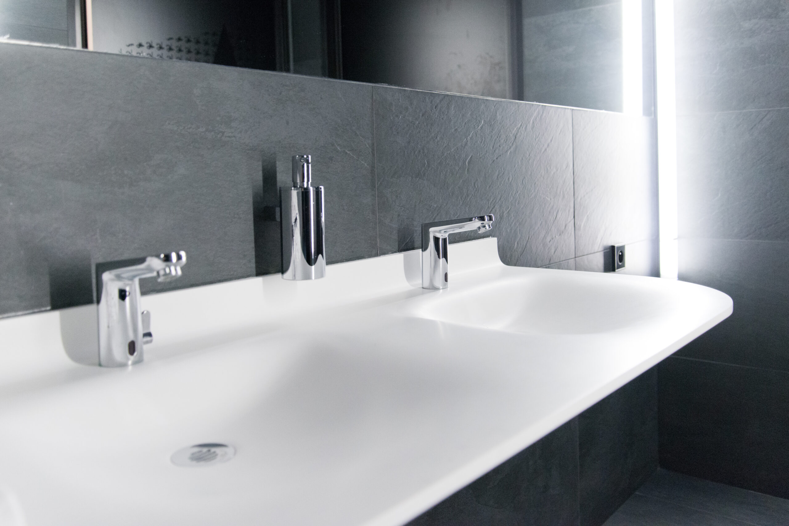 Double vasque d’un seul tenant avec deux robinets distincts, accompagnée d’un grand miroir en façade et d’un éclairage LED intégré sur le côté droit — un agencement moderne et élégant signé Arche du Bois, alliant praticité et design lumineux.