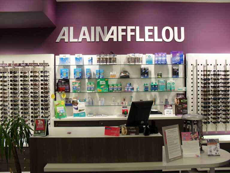 Comptoir d’accueil dans une boutique Alain Afflelou, avec en arrière-plan des étagères murales exposant une large sélection de lunettes — un agencement sobre et professionnel pensé pour une expérience client fluide.