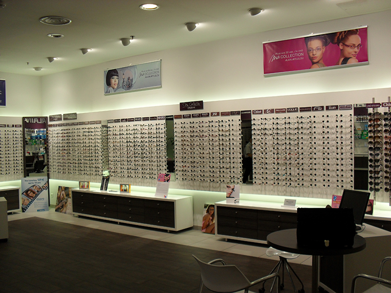 Comptoir d’accueil dans une boutique Alain Afflelou, avec en arrière-plan des étagères murales exposant une large sélection de lunettes — un agencement sobre et professionnel pensé pour une expérience client fluide.