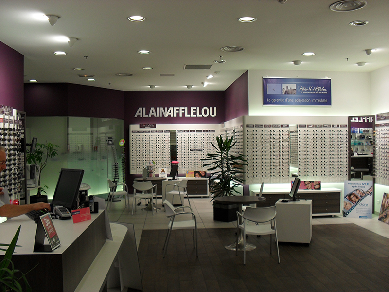 Intérieur d’un magasin d’optique avec tables rondes et chaises pour l’accueil client, entouré de rayonnages exposant des lunettes — un agencement fonctionnel et convivial conçu pour optimiser l’expérience d’achat.