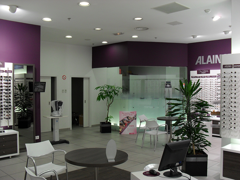 Intérieur d’un magasin d’optique avec tables rondes et chaises pour l’accueil client, entouré de rayonnages exposant des lunettes — un agencement fonctionnel et convivial conçu pour optimiser l’expérience d’achat.