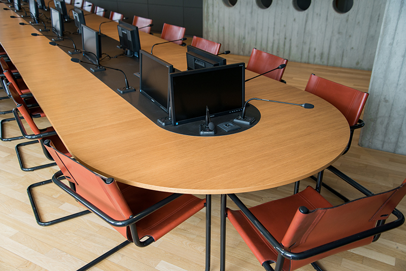 Table de conférence entourée de plusieurs chaises, avec un microphone et un écran individuel devant chaque siège