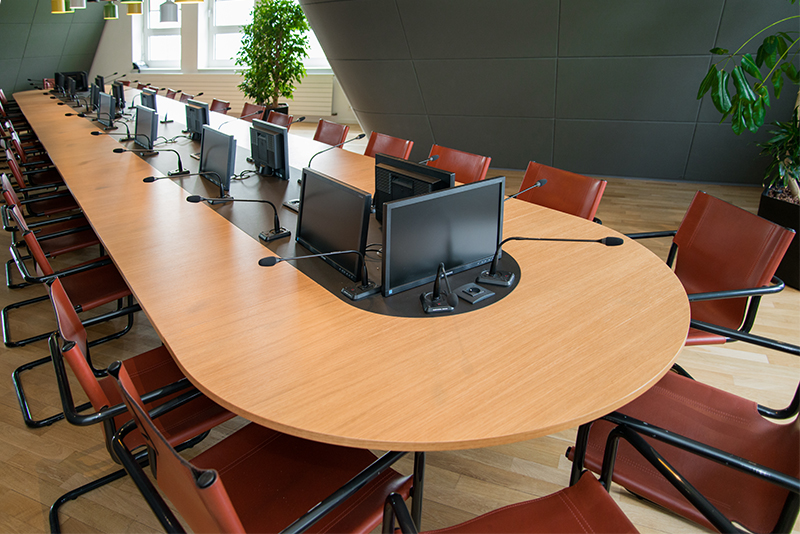 Table de conférence entourée de plusieurs chaises, avec un microphone et un écran individuel devant chaque siège