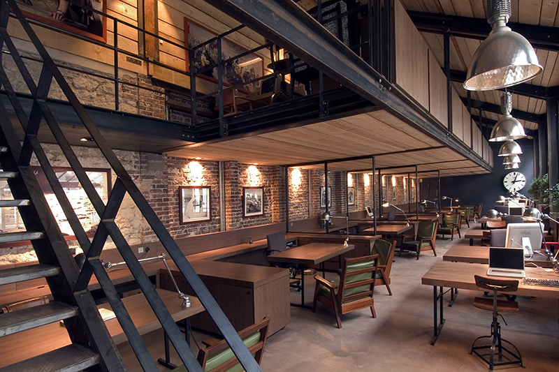 Bureau à l’ambiance boisée avec escalier à gauche, plusieurs tables et chaises modernes en bois, et luminaires suspendus de grande hauteur — un aménagement chaleureux et fonctionnel réalisé par Arche du Bois, combinant design contemporain et matériaux naturels