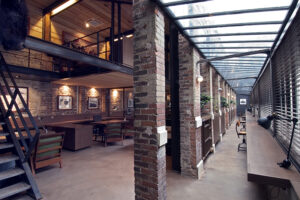 Colonnes en briques apparentes dans une salle spacieuse, avec un couloir sur la droite de l’image et un escalier visible sur la gauche — un agencement architectural soigné par Arche du Bois, mêlant matériaux bruts et circulation fluide dans l’espace. agencement de restaurant sur mesure