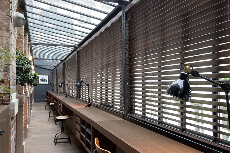 Grandes fenêtres équipées de stores vénitiens avec un long bureau continu placé en façade, chaque poste étant équipé d'une lampe de travail — un aménagement signé Arche du Bois, alliant fonctionnalité, lumière naturelle et design épuré pour un espace de travail optimisé.