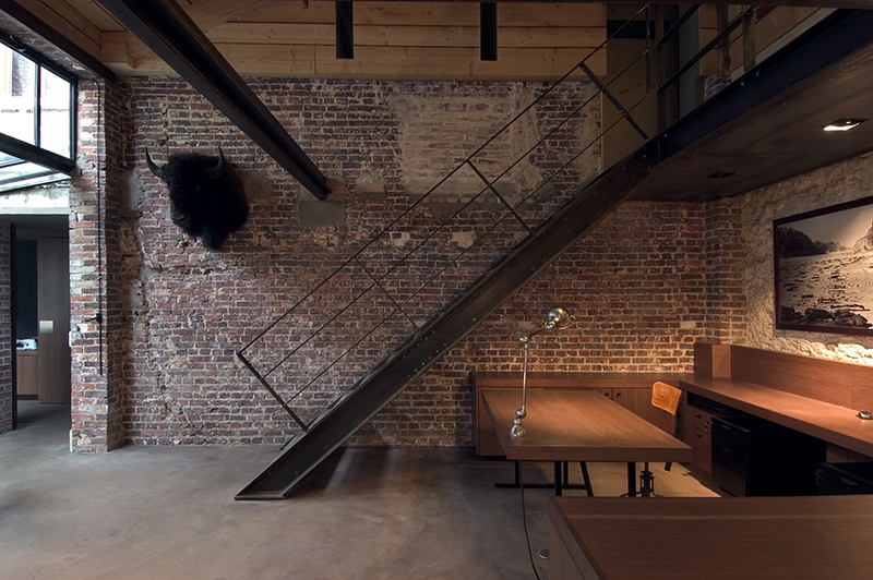 Escalier central avec murs en briques apparentes, tête de taureau naturalisée (taxidermie) accrochée au mur, et sur la droite plusieurs tables et chaises — un espace de caractère agencé par Arche du Bois, combinant matériaux bruts et éléments décoratifs singuliers.