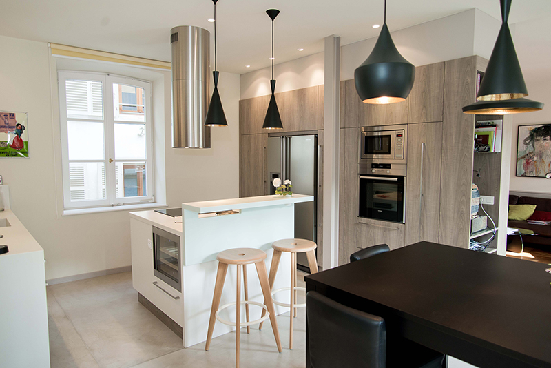 Cuisine sur mesure réalisée par L’Arche du Bois, avec un îlot central, une table brune à droite, quatre suspensions noires au plafond, une grande fenêtre, et au fond des armoires intégrées avec réfrigérateur, micro-ondes et four.
