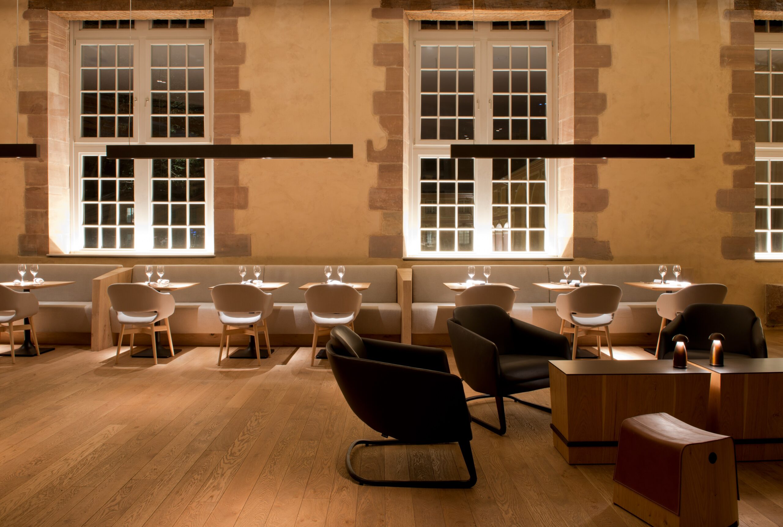 Plusieurs tables et chaises de salle à manger disposées dans la pièce, face à deux grandes fenêtres apportant une lumière naturelle généreuse, avec deux suspensions longues accrochées au plafond — un agencement convivial et lumineux signé Arche du Bois.