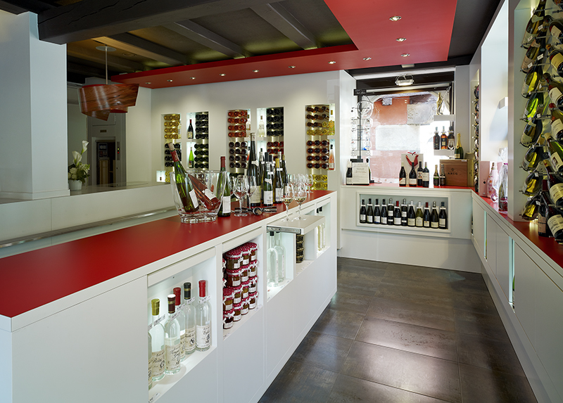 À gauche de l’image, une table avec des bouteilles de vin soigneusement rangées en dessous ; à droite et en face, des étagères remplies de bouteilles — un agencement sur mesure par Arche du Bois, dédié à la mise en valeur et au rangement élégant du vin.