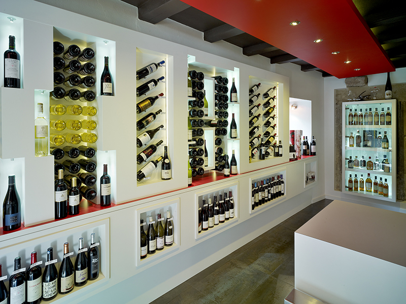 À gauche de l’image, une vitrine expose des bouteilles de vin, tandis qu’à droite se trouve une table en bois, avec en fond un petit réfrigérateur à vin — un agencement raffiné et structuré réalisé par Arche du Bois pour un espace de dégustation ou de présentation.