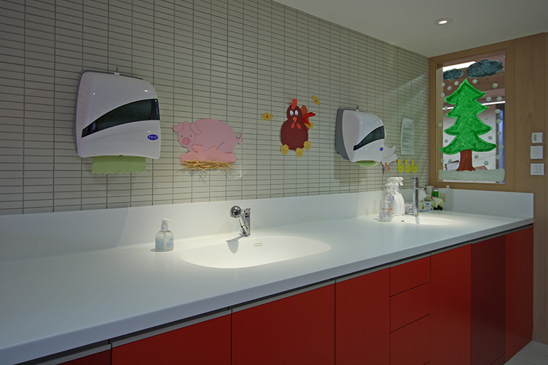 Meuble vasque rouge avec deux lavabos intégrés et deux distributeurs d’essuie-mains installés au-dessus, un agencement sanitaire sur mesure réalisé par Arche du Bois alliant esthétique et praticité.