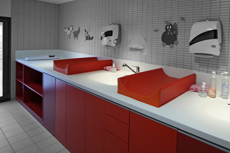 Meuble vasque rouge avec deux lavabos intégrés et deux distributeurs d’essuie-mains installés au-dessus, un agencement sanitaire sur mesure réalisé par Arche du Bois alliant esthétique et praticité.