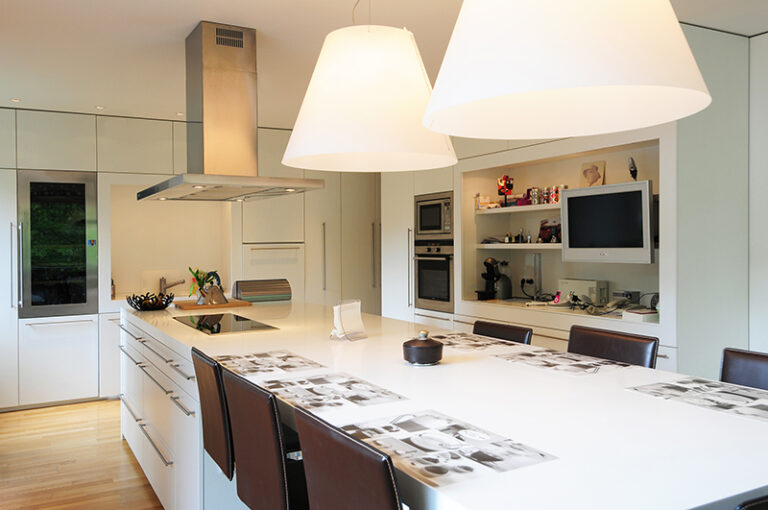 Cuisine sur mesure par LDC Agencement – L’Arche du Bois : table avec tabourets, îlot central avec plaque de cuisson intégrée et hotte, à droite réfrigérateur et placards, ainsi que placards muraux avec étagères, four, micro-ondes et télévision.