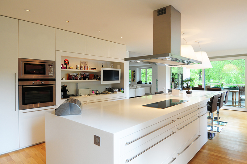 Cuisine sur mesure par LDC Agencement – L’Arche du Bois : table avec tabourets, îlot central avec plaque de cuisson intégrée et hotte, à droite réfrigérateur et placards, ainsi que placards muraux avec étagères, four, micro-ondes et télévision.