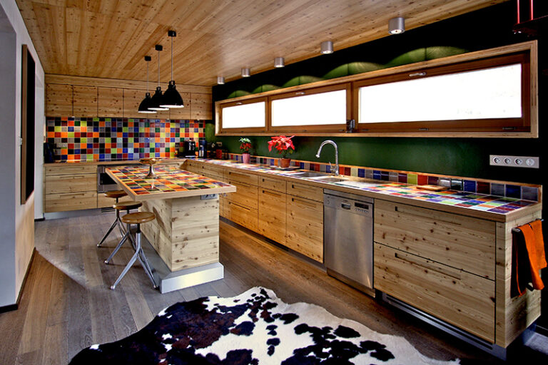 Agencement de cuisine sur mesure par LDC Agencement – L’Arche du Bois : armoires hautes en bois avec plan de travail en carreaux colorés sous trois fenêtres, îlot central en bois, plafond bois et trois suspensions design au-dessus de l’îlot.