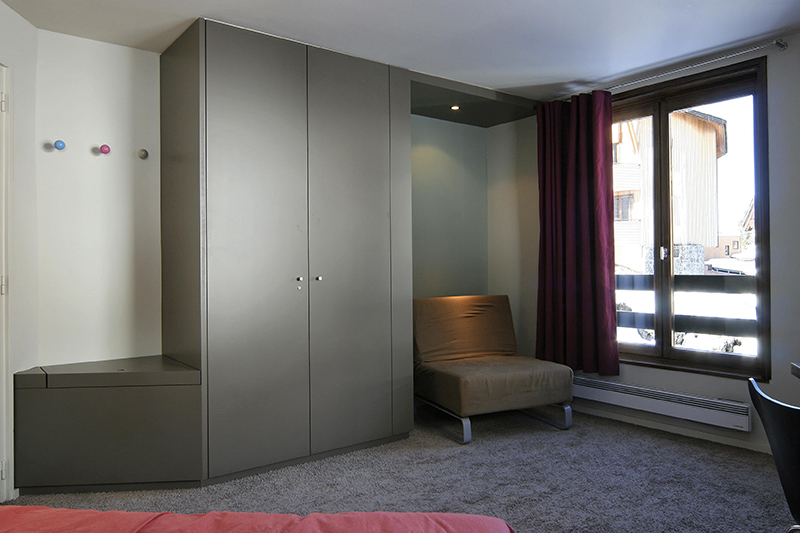 Vue d’une chambre avec une armoire en face, un fauteuil à droite et une fenêtre attenante apportant de la lumière naturelle.