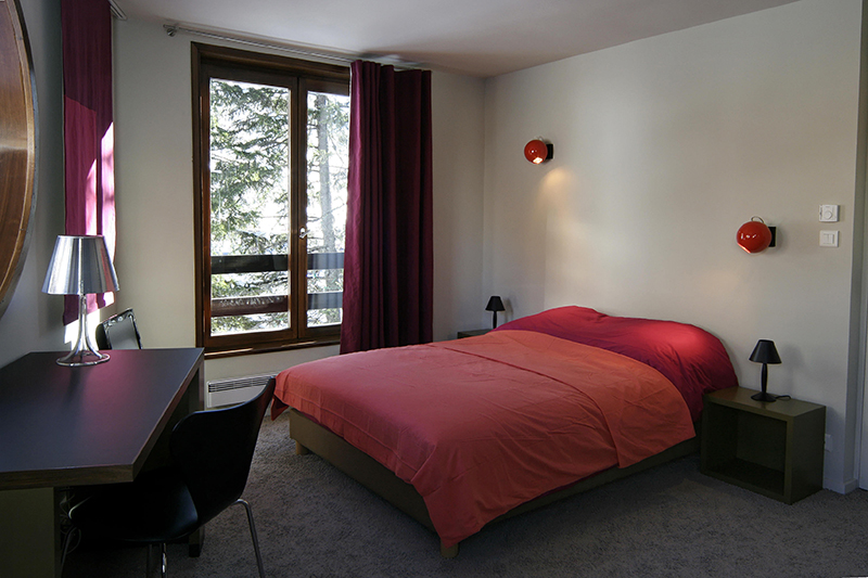 Chambre à coucher avec un lit rouge au centre, deux lampes au-dessus du lit, un bureau avec chaise à gauche et une grande fenêtre en face laissant entrer la lumière naturelle.