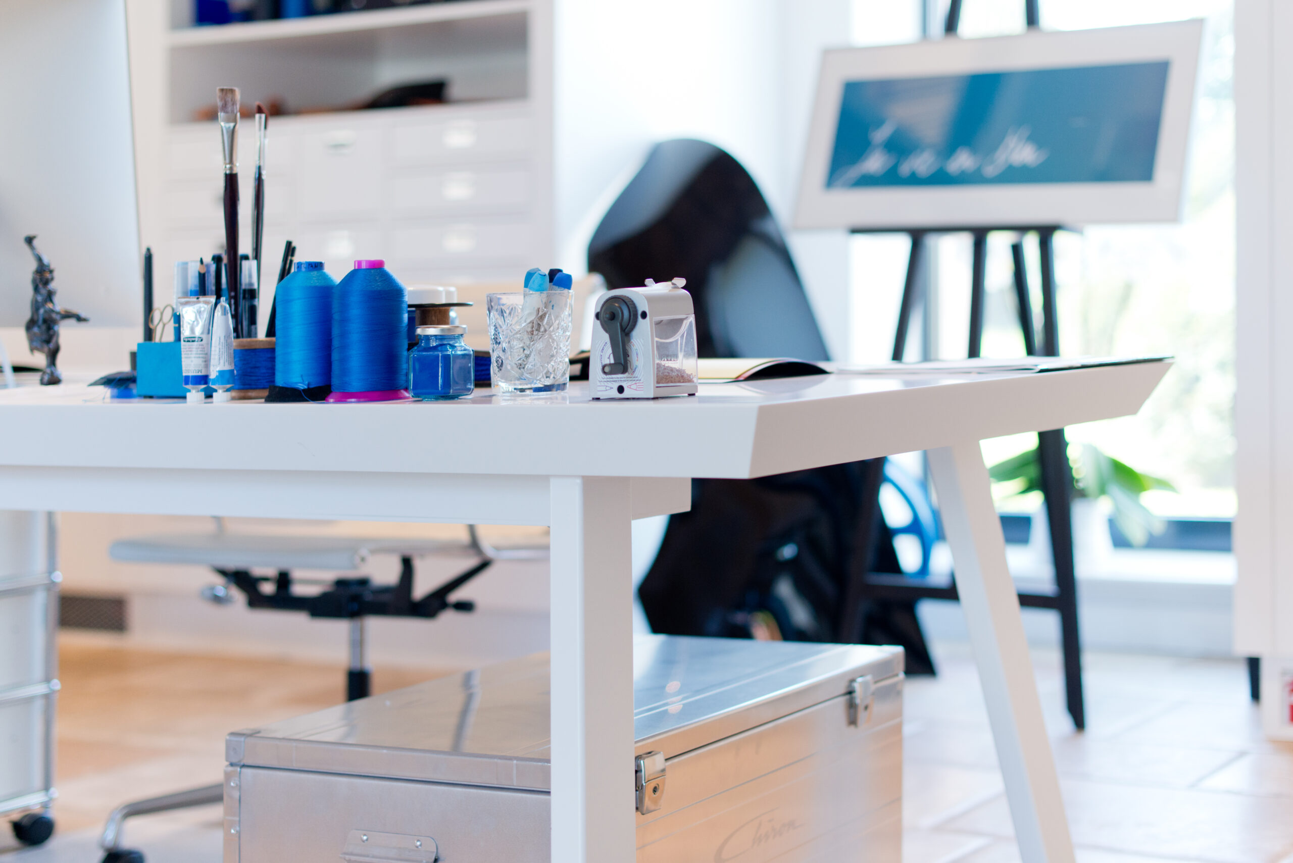 Bureau blanc aux pieds inclinés, accompagné à l’arrière d’un chevalet avec toile ; sur le plateau, des fournitures artistiques telles que taille-crayon, pinceaux et matériel de dessin, et un coffre de rangement est placé sous le bureau