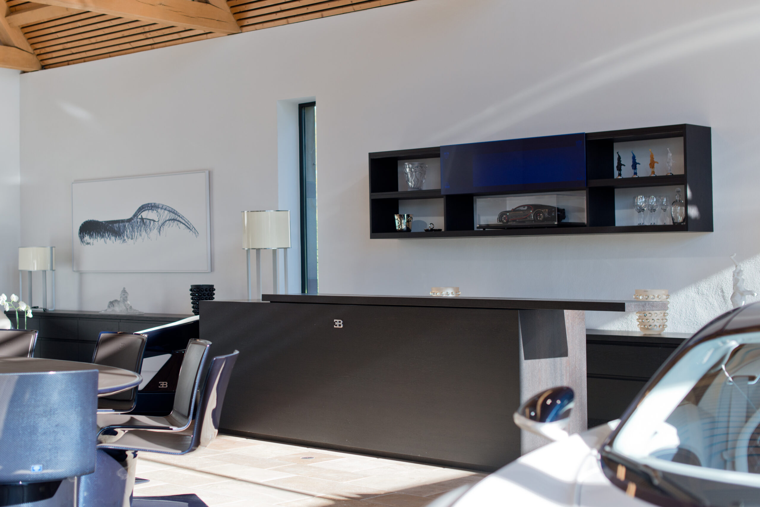 Espace d’accueil de l’entreprise Bugatti avec comptoir noir arborant le logo de la marque, surmonté d’une étagère murale contenant des objets décoratifs en verre et une maquette de voiture Bugatti ; à droite, l’avant d’un véhicule exposé, et à gauche, une table ronde entourée de chaises ainsi qu’un cadre blanc accroché au mur — un aménagement d’exception signé Arche du Bois, alliant prestige automobile et design intérieur sur mesure