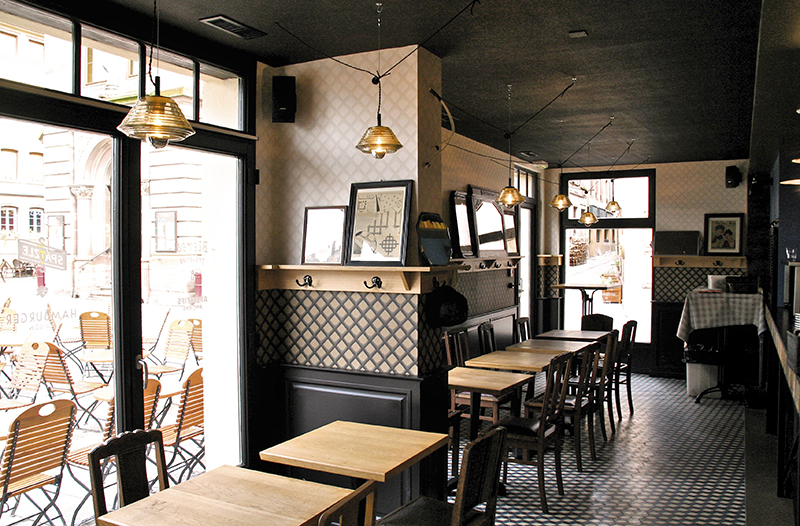 À droite de l’image, une porte vitrée donnant sur l’extérieur, avec une rangée de tables et chaises en bois alignées devant ; à l’arrière-plan, une grande fenêtre en verre apportant luminosité et ouverture — un agencement naturel et fonctionnel signé Arche du Bois.