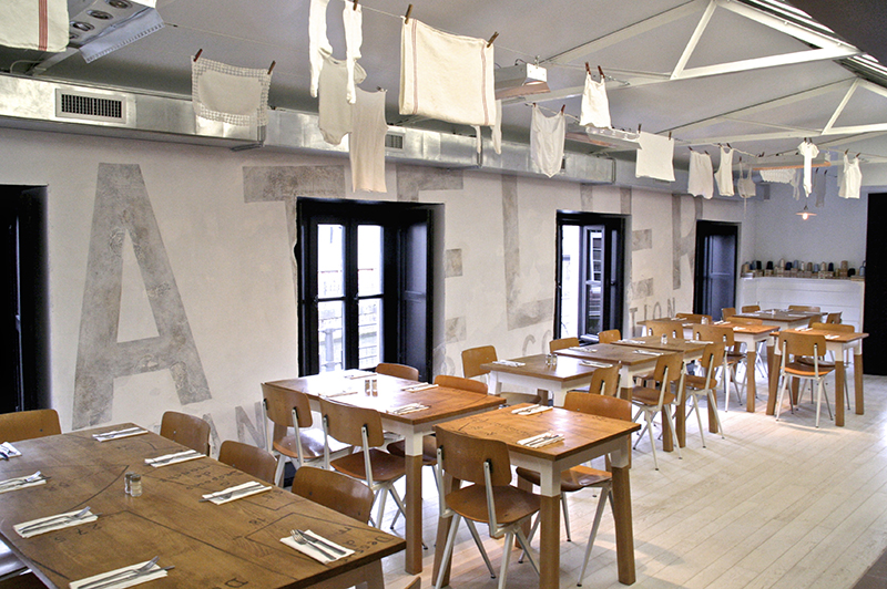 Plusieurs tables et chaises en bois disposées dans la pièce, face à trois grandes fenêtres laissant entrer la lumière naturelle — un agencement lumineux et convivial réalisé par Arche du Bois, alliant confort et matériaux naturels.