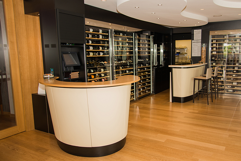 Espace accueil courbe situé à gauche de l’image, accompagné d’une vitrine à portes vitrées sur la droite, utilisée pour le stockage et l’exposition de bouteilles de vin — un agencement sur mesure signé Arche du Bois, mêlant élégance et fonctionnalité.