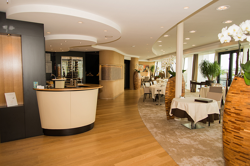 Salle à manger avec, sur la gauche, un comptoir d’accueil en bois, et sur la droite, un ensemble de tables et chaises — un agencement intérieur signé Arche du Bois, marqué par l’utilisation de formes courbes et d’un design fluide et chaleureux.