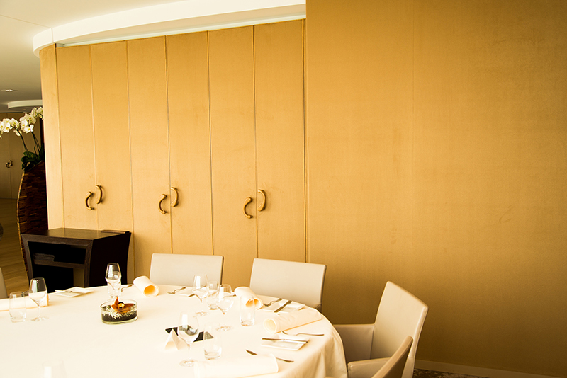 Moitié d’une grande table ronde visible au premier plan, avec en arrière-plan une armoire en bois clair — un agencement sobre et fonctionnel conçu par Arche du Bois, alliant convivialité et rangement discret.