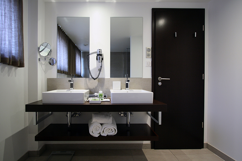 Deux vasques indépendantes avec leurs miroirs respectifs fixés au mur en face, et une porte visible sur la droite de l’image — un agencement fonctionnel et raffiné signé Arche du Bois pour un espace sanitaire contemporain.