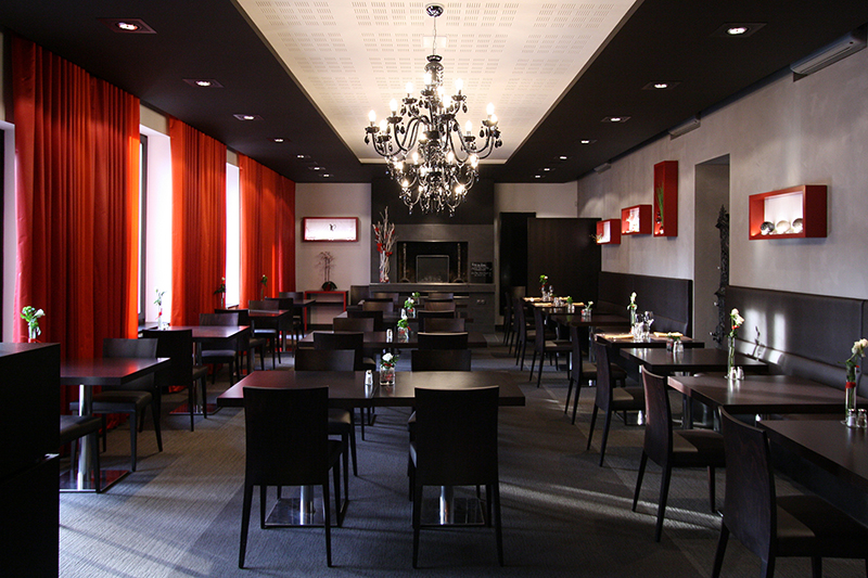 Grande salle à manger aménagée avec de nombreuses tables et chaises, éclairée naturellement grâce à des fenêtres habillées de rideaux sur le côté gauche — un agencement spacieux, chaleureux et fonctionnel signé Arche du Bois.