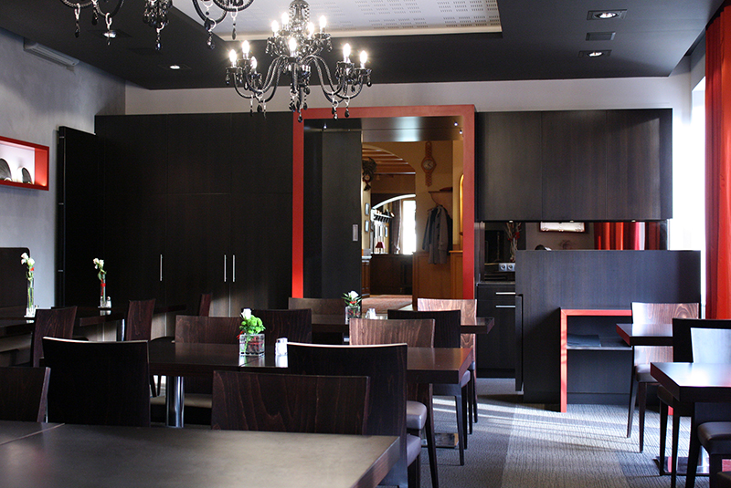 Salle à manger avec plusieurs tables et chaises, donnant sur une entrée visible en fond de pièce — un agencement fluide et convivial réalisé par Arche du Bois, pensé pour le confort et la circulation naturelle des usagers.