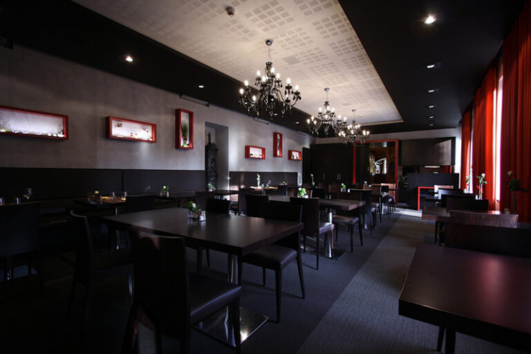 Salle à manger spacieuse aménagée avec de nombreuses tables et chaises, éclairée par des fenêtres avec rideaux sur la droite, et décorée de cadres muraux sur la gauche — un agencement chaleureux et équilibré signé Arche du Bois.