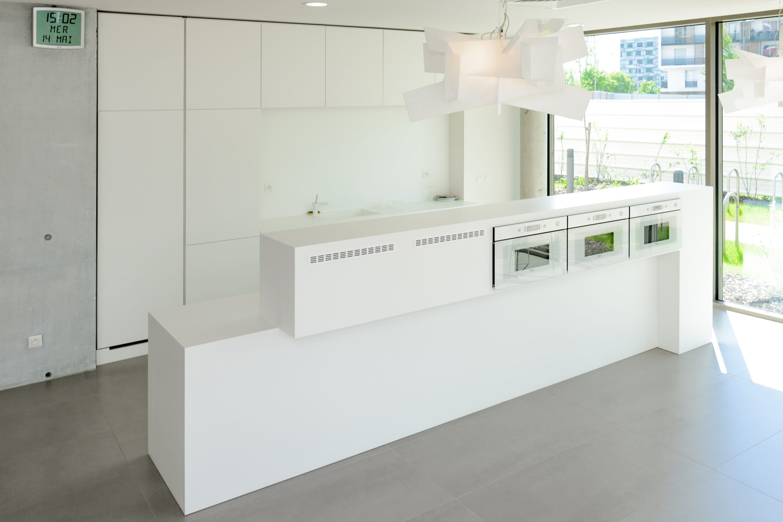 Grand plan de travail blanc avec trois micro-ondes posés dessus, éclairé par une suspension centrale ; à droite, une porte vitrée, et à l’arrière, plusieurs armoires de rangement — un aménagement pratique et épuré réalisé par Arche du Bois.