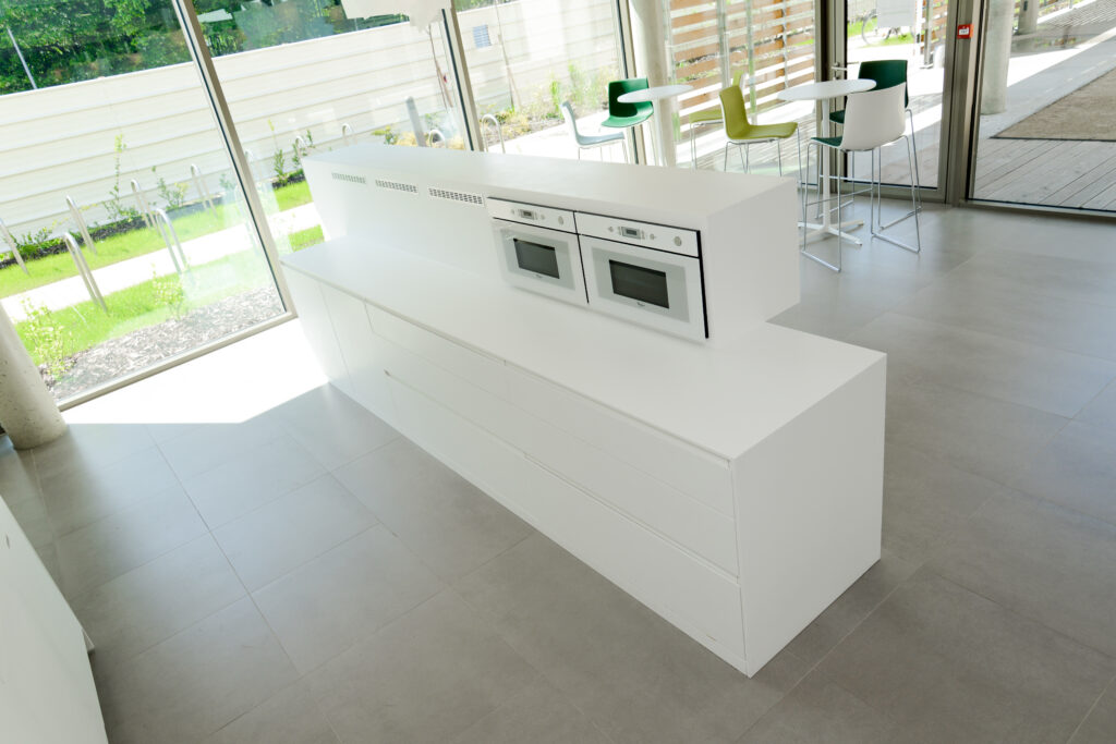Cuisine sur mesure par LDC Agencement – L’Arche du Bois : îlot central blanc avec four et micro-ondes intégrés, tiroirs en bas, entouré de portes et fenêtres, avec deux petites tables et six tabourets.