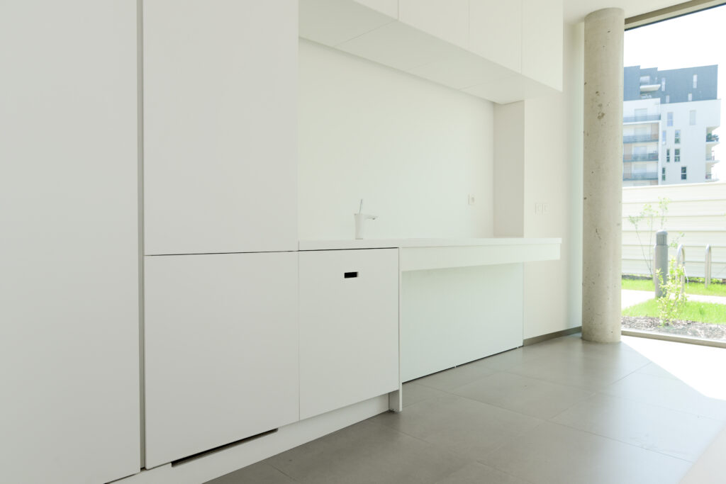 cuisine sur mesure par LDC Agencement – L’Arche du Bois : à gauche meubles de rangement et placards blancs avec robinet, à droite porte vitrée et colonne.