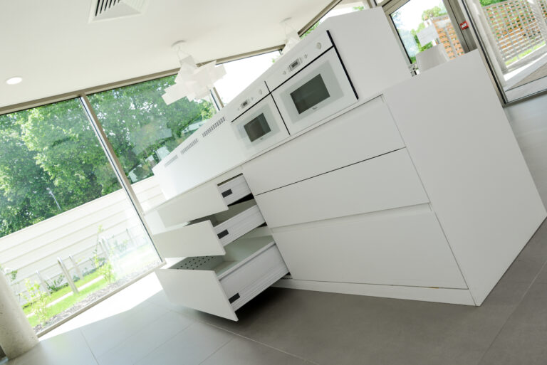 Cuisine sur mesure par LDC Agencement – L’Arche du Bois : tiroirs ouverts à gauche et fermés à droite, murs vitrés tout autour, avec micro-ondes et four intégrés dans les placards.