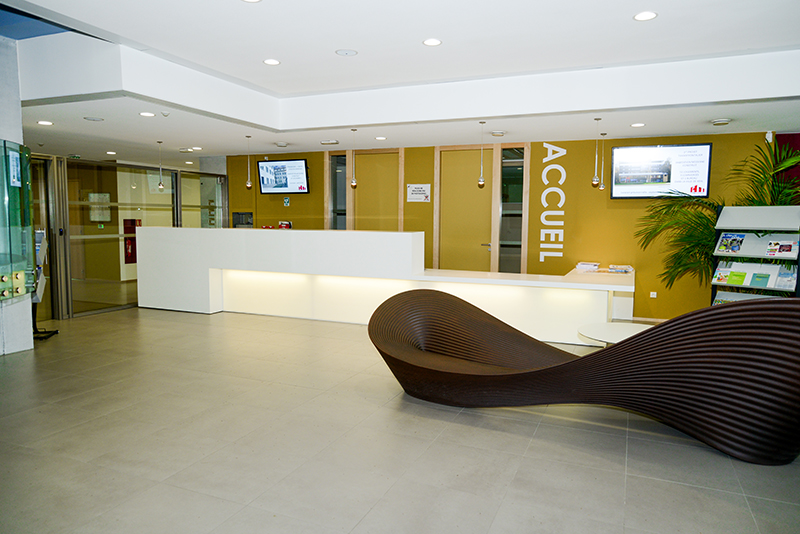 Comptoir d’accueil blanc avec éclairage LED intégré en dessous, placé devant un mur jaune vif équipé de deux écrans ; sur la droite, un fauteuil ergonomique au design singulier complète cet espace contemporain conçu par Arche du Bois