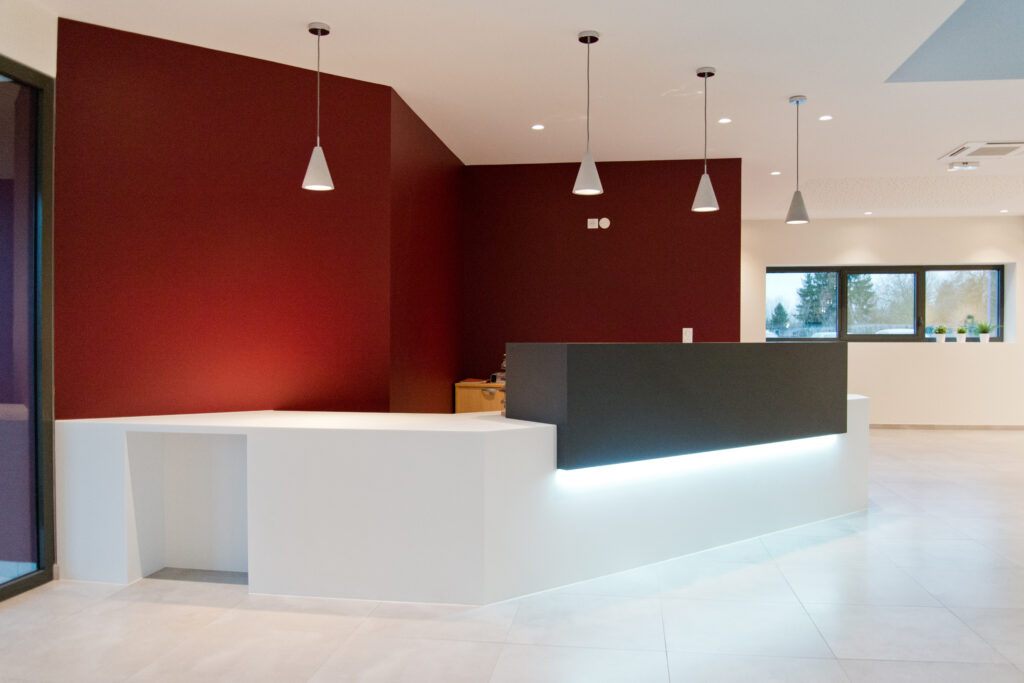 Comptoir d’accueil blanc avec bandeau supérieur noir et éclairage LED intégré en dessous, installé devant un mur rouge ; quatre suspensions lumineuses au plafond et une fenêtre en fond à gauche complètent l’agencement — une réalisation contemporaine et soignée signée Arche du Bois, combinant contrastes de couleurs et mise en lumière efficace.