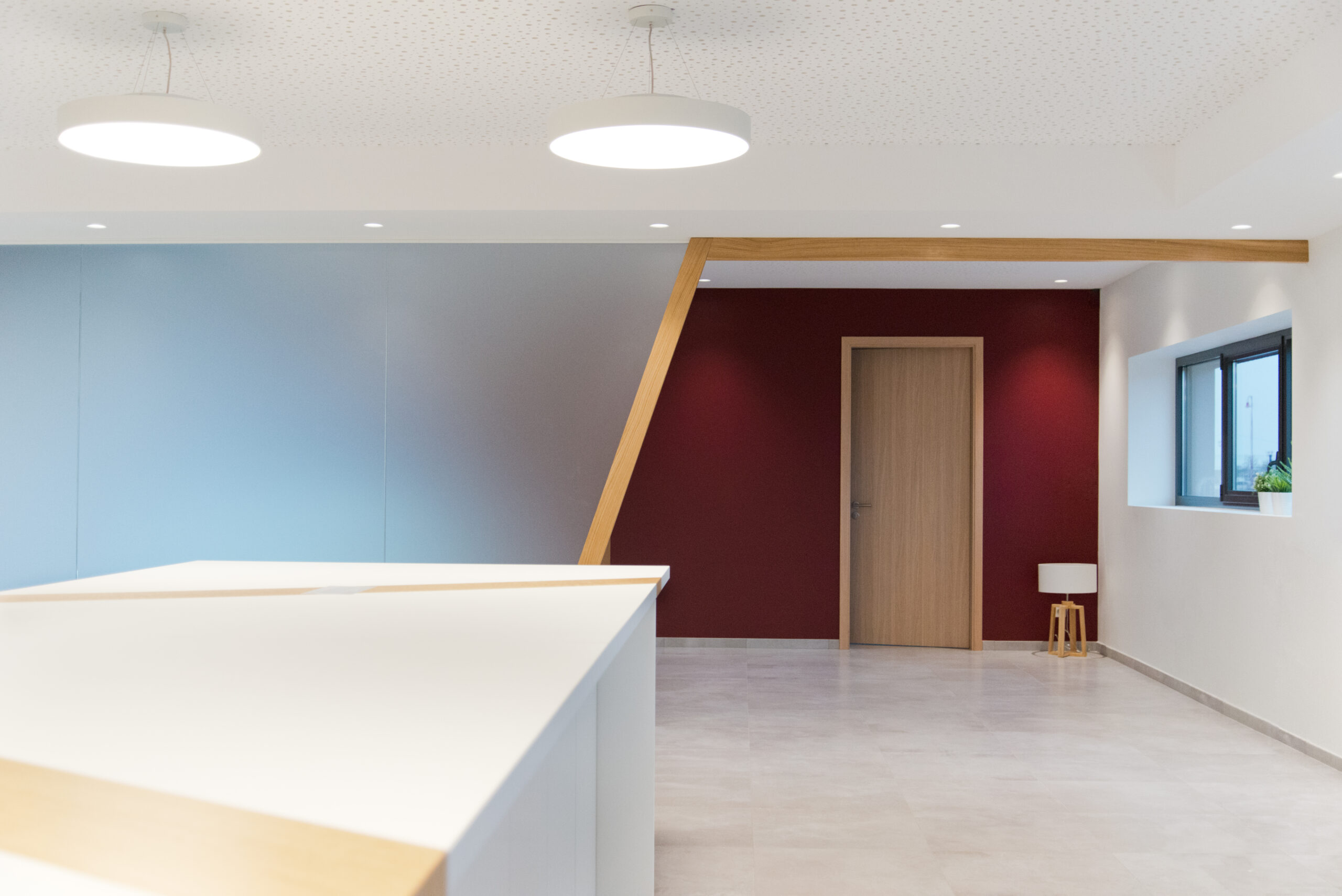 Vue d’un couloir ou d’un salon avec une porte au fond, encadrée par un mur rouge contrastant avec le mur latéral droit de couleur blanche, où se trouve également une fenêtre