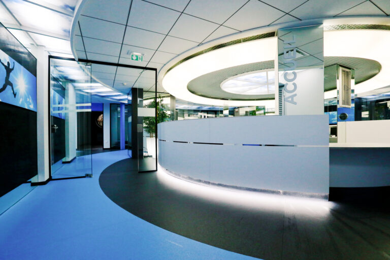 Espace accueil circulaire conçu par Arche du Bois, avec un miroir mural en face, une porte vitrée sur la gauche, et un luminaire LED circulaire au plafond diffusant des teintes bleues et blanches — un agencement immersif et contemporain.